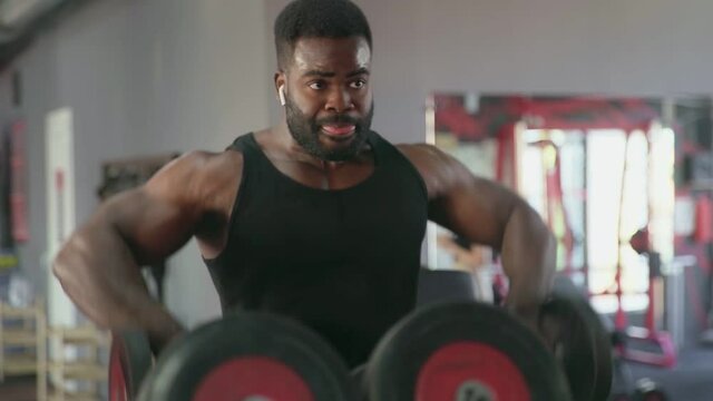 African american man bodybuilder with wireless headphones performing Side raise exercise with dumbbells at gym