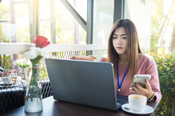 Asian woman using laptop computer and smart phone with white coffee cup on table in cafe.