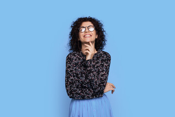 Fototapeta premium Adorable woman with curly hair and glasses is smiling at camera on a blue studio wall