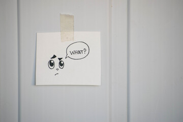Sign "What?" with a cute face is attached to a white wall with tape