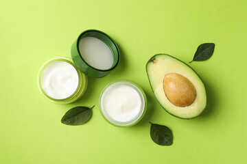 Jars of cosmetic cream, leaves and avocado on green background