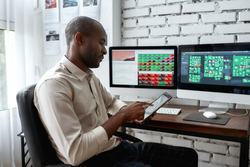 Fototapeta na wymiar For the accessible possibility. Stylish african businessman, trader sitting by desk in front of multiple monitors and using his smartphone to check charts while working in the office.