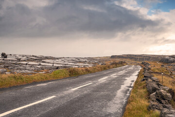 Small narrow asphalt road to a mountain. Burren region, county Clare, Ireland. Low cloudy sky. Nobody,