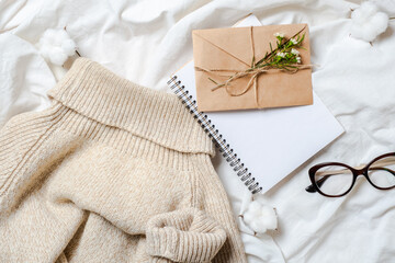 Romance flat lay composition with blank paper notepad, craft paper envelope with flowers, woolen sweater, glasses, cotton on girl's bed. Top view festive feminine accessories. Christmas letter concept