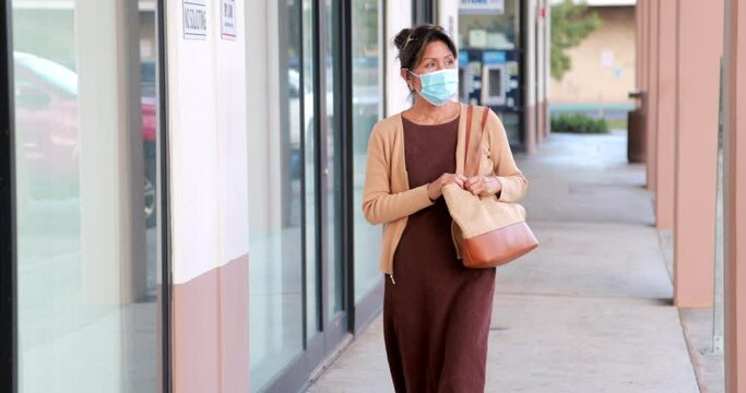 Attractive Professional Woman Dressed In Brown Casual Wearing Covid Mask For Coronavirus Pandemic Walking Along Of Roll Of Glass Windows Outside Stops, Pulls Cell Phone From Purse And Begins Texting  