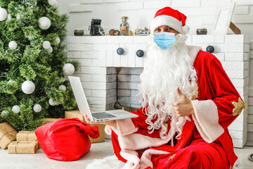 Obraz premium Santa Claus wearing face mask, holding gift box on xmas eve sitting at cozy home using laptop computer. Merry Christmas Covid 19 coronavirus social distance concept.