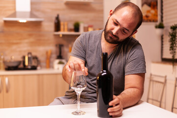 Alcoholic man being depressed and frustrated holding bottle of red wine in kitchen. Unhappy person disease and anxiety feeling exhausted with having alcoholism problems.