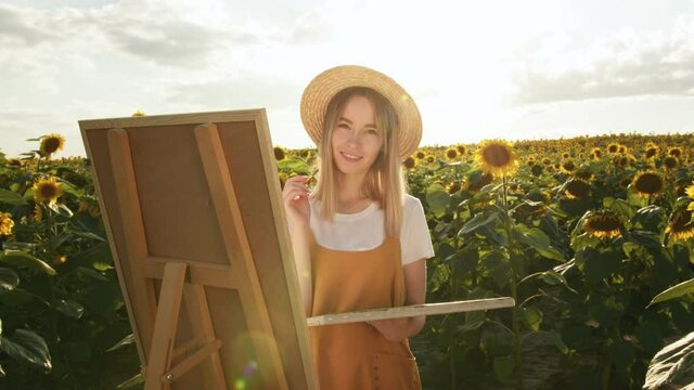 A woman is standing in a field of sunflowers and drawing a picture. She is looking at the camera and smiling. 4K