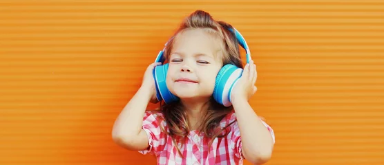 Poster Portrait of little girl child in wireless headphones listening to music over orange background © rohappy