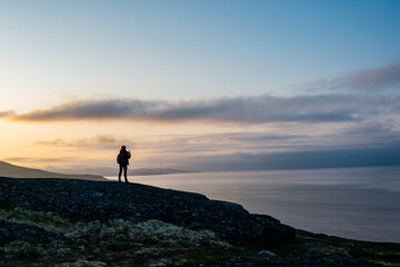 Silhouette figure of young woman traveller enjoying the Beaty of nordic nature on the mountain near the sea taking the picture of the sunset 
