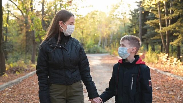 Woman and her son at an empty autumn park wearing face surgical masks for protection against virus. Mom takes son's hand and they look at each other with love. New normality video outdoors. Covid-19