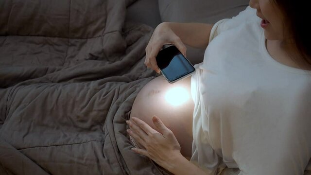 asian pregnant woman playing with baby in belly with flashlight from mobile smart phone on bed in bedroom. pregnancy female touching abdomen.
