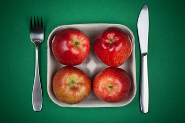Top view of four fresh red apples in a pulp paper tray. Cutlery, fork and knife, on the sides. Studio photo isolated on green background.