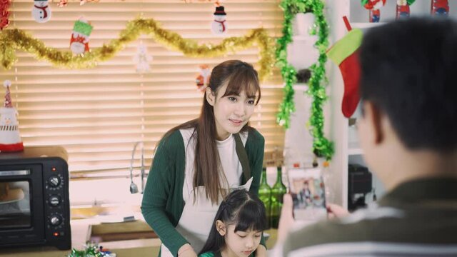 Young beautiful Asian mother and lovely daughter looking and smiling at the camera from the smartphone taken by father while making Christmas cake together in Christmas decorated kitchen at home.
