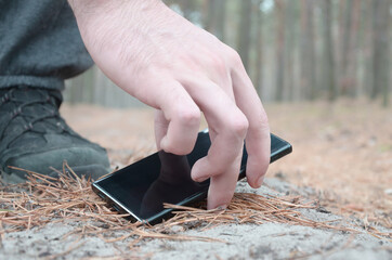 Male hand picking up lost mobile phone from a ground in autumn fir wood path. The concept of...