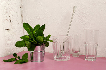 Closeup of bucket full of fresh mint leaves, empty glasses, spoon on the pink bar counter against white wall