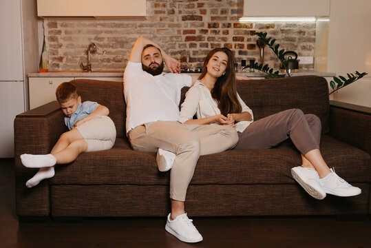 Dad And Mom Are Watching TV On The Sofa While The Son Is Sleeping Near In The Evening. Family At Home.