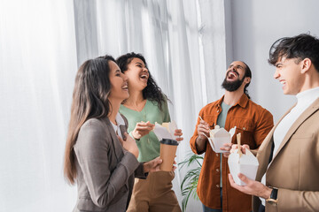 excited bearded businessman laughing near hispanic colleagues holding thai noodles and coffee to go
