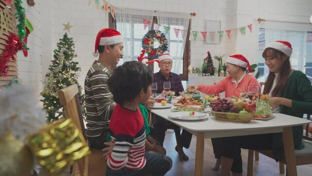 Happy Asian Family, Grandparents, Father, Mother, Lovely Daughter And Little Son Enjoying Christmas Dinner Together In Christmas Decorated Dining Room At Home And Father Gave Mother A Food On Plate.