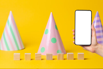 Paper multicolored holiday caps on a yellow background. Wooden cubes for writing. A woman's hand holds a smartphone in her hand. Mock up. The concept of party and online communication