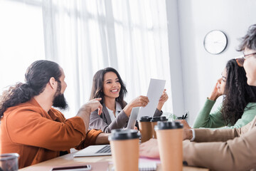 Hispanic office worker pointing with finger at document in hands of smiling co-worker in office on blurred foreground