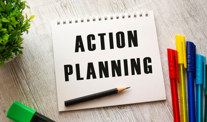 A notebook on a spring with the text ACTION PLANNING lies on a wooden table with colored pens.