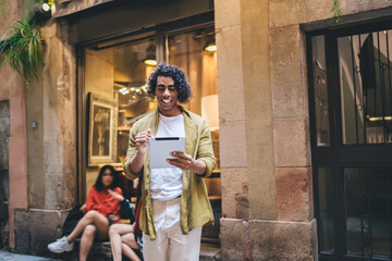 Cheerful curly hipster guy in casual wear walking on city street checking notification on touchpad enjoying free time, smiling handsome man blogger sending text messages and chatting outdoors