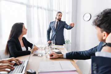 Confident hispanic executive pointing with finger at co-worker during business meeting in office on blurred foreground
