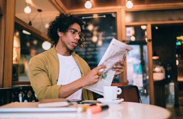 Serious handsome male 20s in trendy wear and eyeglasses enjoying leisure holding newspaper getting daily information, pensive businessman checking financial economy news from columns in print media