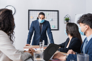 Hispanic businessman in medical mask looking at colleagues during meeting in office