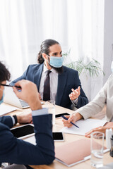Hispanic businessman in medical mask working near colleagues and devices on blurred foreground