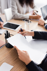 Cropped view of businesswoman working with documents near colleagues and devices on blurred background
