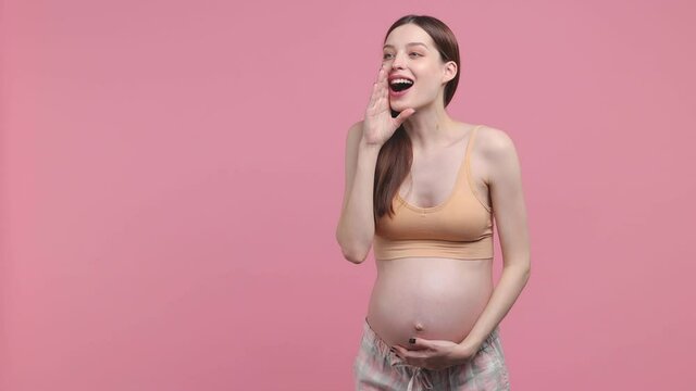 Fun young pregnant woman future mom in basic top stroking keeping hands on belly tummy with baby scream aside isolated on pastel pink background studio. Maternity family pregnancy gynecology concept