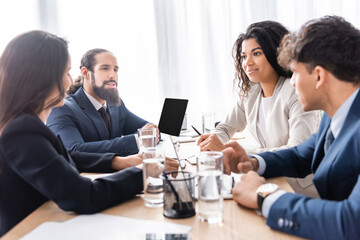Smiling hispanic businesswoman working with colleagues near laptop and water on blurred foreground on table