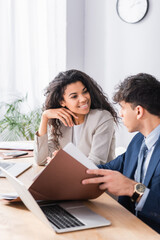 Hispanic businesswoman smiling at colleague with papers near laptop on blurred foreground