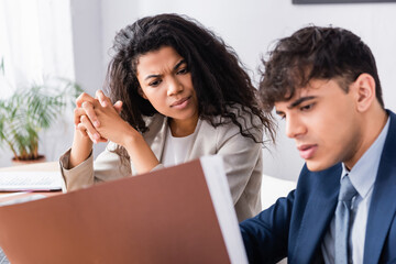 Obraz premium Pensive hispanic businesswoman looking at papers near colleague on blurred foreground in office