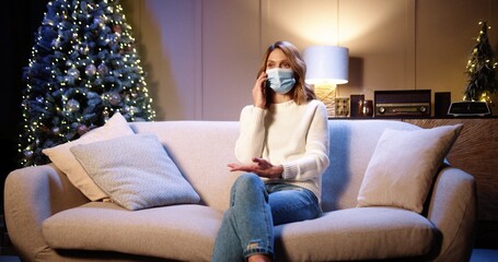 Obraz premium Happy Caucasian beautiful female in medical mask sitting on sofa at decorated home near xmas glowing tree in evening and calling on smartphone greeting friend with holidays. Christmas concept