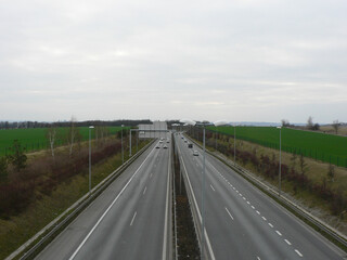 Fototapeta premium Empty highway on a cloudy day. A speeding car. View over the two-way road with two lanes in each direction. Green field along the road, autobahn or highway.