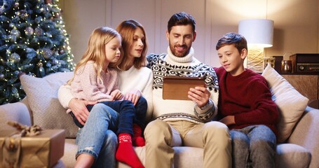 Happy Caucasian family with kids sitting in living cozy room with glowing xmas tree and looking at tablet choosing gifts Parents talking with children on new year eve. Happy holiday concept