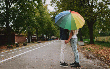 Gay couple is standing under the umbrella and holding their hands. Two men lovers in the park. Colorful umbrella like symbol of LGBT community. Equal rights