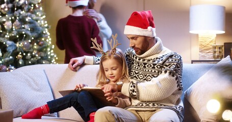 Portrait of joyful caring Caucasian dad with little girl sitting in cozy room at home and reading book. Mom and son decorating glowing Christmas tree. Family holiday time concept. New Year celebration