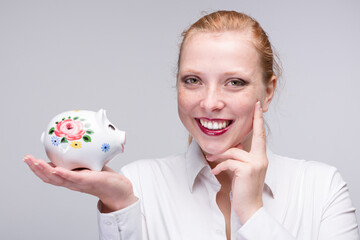Fototapeta na wymiar Young happy red haired woman presenting a piggy bank / porcelain bank in her hand