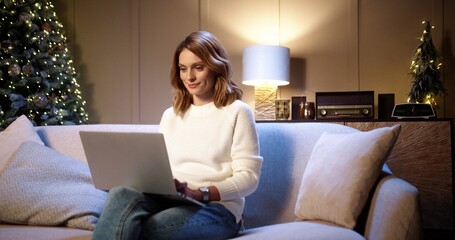 Happy young beautiful Caucasian woman sitting on sofa at decorated home near glowing christmas tree...