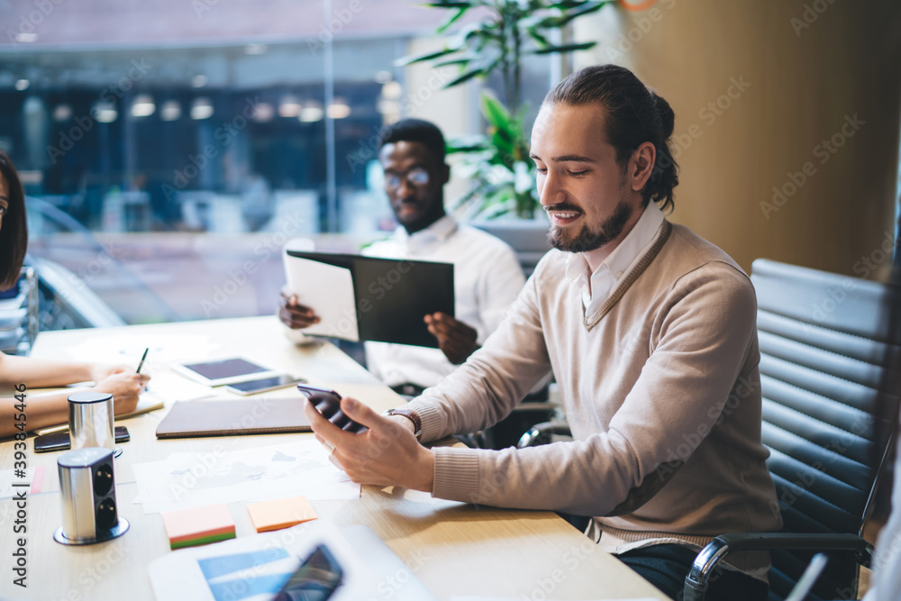 Poster cheerful caucasian male employee in smart casual wear sitting at working place with colleagues check