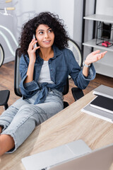 joyful young woman talking on smartphone near laptop on blurred foreground