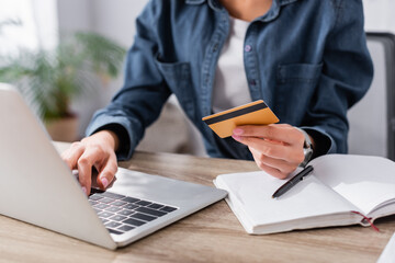 partial view of young woman holding credit card near laptop on desk