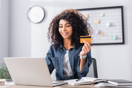 cheerful young woman holding credit card near laptop on desk