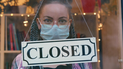 Young woman employee in safrty mask and glasses turning closed sign on shop door
