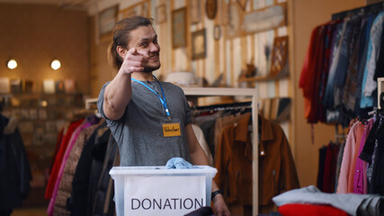 Young man showing volunteer badge and pointing fingers at camera in charity shop