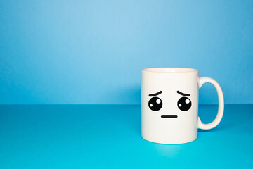 Close-up of a white mug on a blue background looking sad. Concept of sadness caused by Covid-19, new normal and Blue Monday.